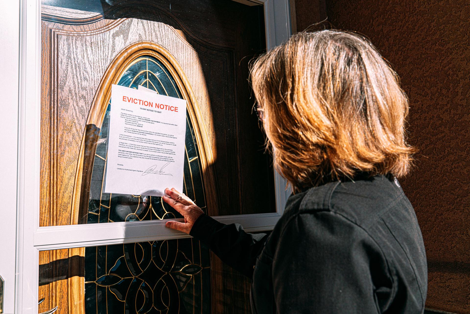 Middle-Age Woman Reaching and Removing an Eviction Notice Taped on the Front Door of a Home in a Residential Suburban Neighborhood